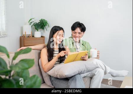 Una bella giovane coppia asiatica si diverte a conversare leggendo un libro insieme sul divano del soggiorno. rapporto sano, legame, domesti Foto Stock