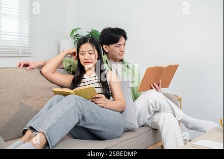 Una giovane coppia di sposi asiatica felice e accogliente si diverte a casa, leggendo libri sul divano al mattino. nuova coppia sposata, sano r Foto Stock