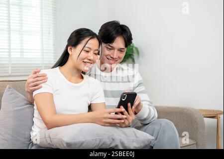 Una giovane e felice coppia asiatica si siede insieme su un divano nel soggiorno, guardando un video su uno smartphone. persone, tecnologia wireless, stile di vita Foto Stock