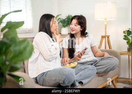 Due giovani amiche asiatiche si siedono su un divano in un salotto minimalista, chiacchierano qualcosa sul loro smartphone, gustando spuntini e shar Foto Stock