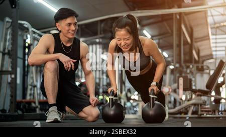Una determinata cliente asiatica viene addestrata da un allenatore di fitness professionista in palestra, eseguendo push-up con kettlebell come parte del suo lavoro Foto Stock