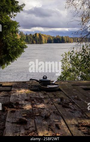 Un momento tranquillo in riva al fiume: Riflessi della foresta, un ponte in legno e utensili da cucina dimenticati che suggeriscono una storia lasciata nell'abbraccio della natura Foto Stock