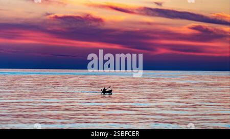Un tramonto sereno sulle acque calme, caratterizzato da un kayak solitario con vivaci sfumature di viola, arancione e rosa nel cielo. Foto Stock
