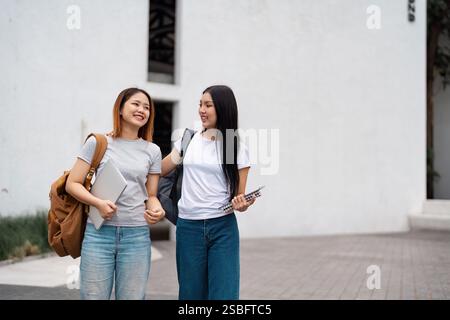 Due diversi studenti universitari che camminano insieme, sorridono e condividono un momento di sostegno nel campus con i loro materiali di studio. Foto Stock