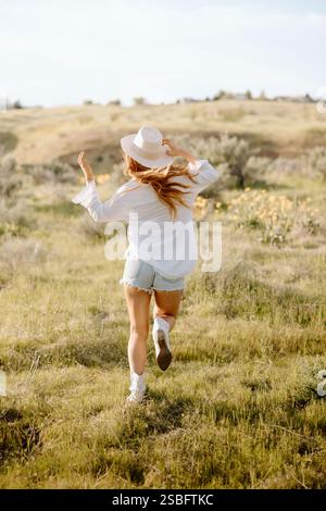 La giovane donna rossa in cappello corre attraverso un campo estivo illuminato dal sole Foto Stock