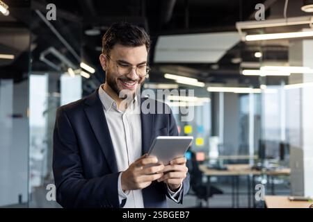 Uomo d'affari di successo in tuta da lavoro con tablet. Uomo all'interno dell'ufficio sul posto di lavoro sorridente felicemente, utilizzando l'app di investimento finanziario. Foto Stock