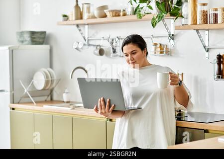 Una giovane donna di grandi dimensioni ama il suo notebook mentre sorseggia una tazza calda in una cucina elegante. Foto Stock