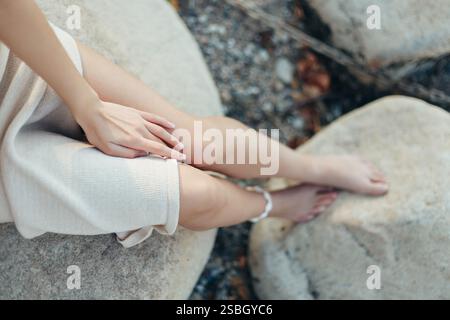 Belle gambe femminili sottili. Una donna a piedi nudi siede sulla pietra in spiaggia con un vestito, vista dall'alto Foto Stock