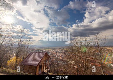 Una vista panoramica in cima alla collina caratterizzata da un'accogliente cabina tra alberi aridi con una vasta città sottostante, incorniciata da un cielo blu nuvoloso. La scena cattura la tranquillità a Foto Stock
