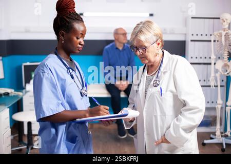 Infermiera afroamericana tiene appunti recensioni note mediche con medico senior, in piedi in stanza clinica. Personale ospedaliero femminile diversificato che discute come vecchio paziente maschio aspetta in background. Foto Stock