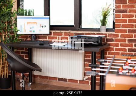 Moderno ufficio di avvio con grafici finanziari sullo schermo del computer, stampante e appunti sul tavolo. Pareti in mattoni luminosi con decorazione vegetale e mobili ordinati vicino alla finestra. Foto Stock