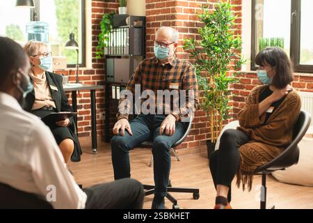 Giovani e anziani seduti in cerchio, partecipano al programma di salute mentale. L'uomo anziano condivide nervosamente la sua sofferenza mentre la psichiatra anziana ascolta attentamente durante la sessione di terapia di gruppo Foto Stock