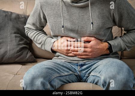 Primo piano di un uomo seduto su un divano a casa, che stringe lo stomaco, soffre di dolore addominale o crampi. Persona che soffre di problemi di salute, diges Foto Stock