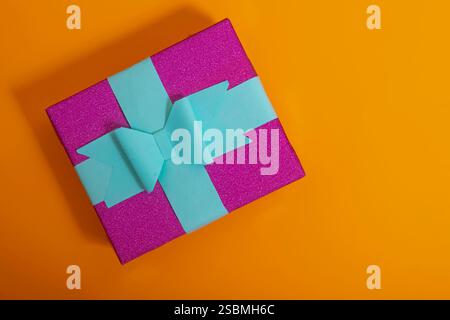 Vista dall'alto di una confezione regalo splendidamente confezionata con carta rosa scintillante e un nastro blu, su un audace sfondo arancione, che evoca festività e celebrazioni Foto Stock