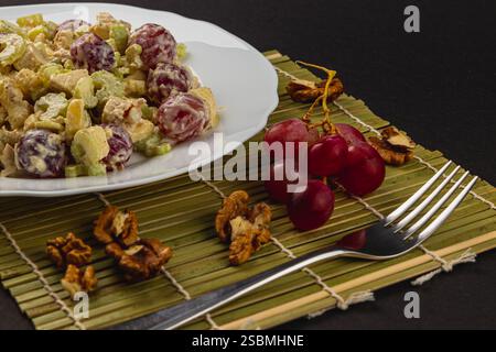Un piatto vivace presenta un mix di filetto di pollo, uva, gambi di sedano e yogurt greco, splendidamente disposto su un piatto bianco sopra un tappetino di bambù. Foto Stock