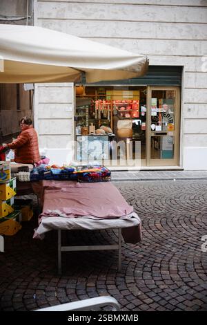 Busseto, Parma, Italia - 31 gennaio 2025 Venditore prepara stand di abbigliamento a Busseto Street Market, Parma, Italia, con negozio di gastronomia sullo sfondo Foto Stock