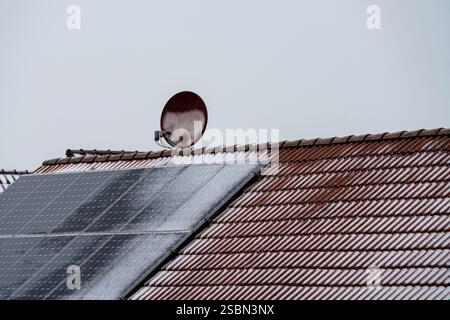 La neve si accumula sui pannelli solari installati su un tetto con tegole rosse, con una parabola satellitare visibile nelle vicinanze. Il cielo è grigio, a indicare il freddo inverno. Foto Stock