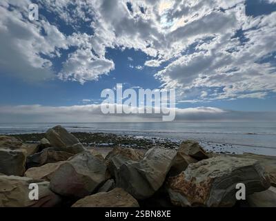 Onde dolci si infrangono su una costa rocciosa dove i massi sono sparsi. Un bellissimo cielo blu è punteggiato da soffici nuvole, creando una tranquilla costa Foto Stock