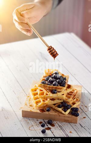 Versare a mano il miele con un cucchiaio di miele di legno su una pila di waffle belgi con mirtilli al sole del mattino Foto Stock