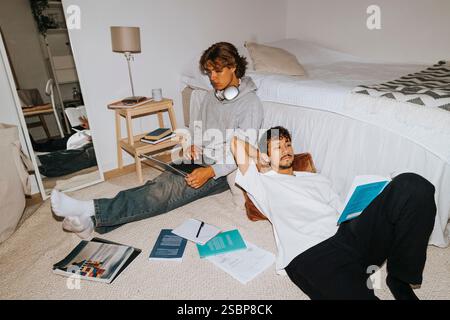Un giovane che legge un libro mentre si sdraia vicino ad un amico maschio usando un portatile nella stanza del dormitorio del college Foto Stock