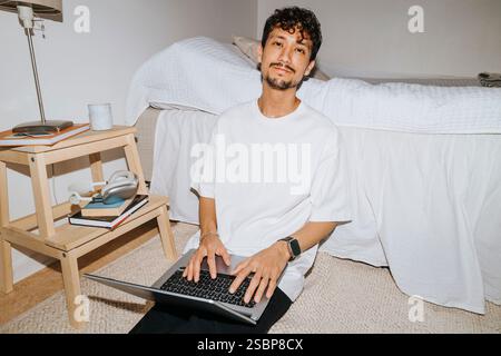 Ritratto di un giovane sorridente seduto con un laptop a letto nel dormitorio del college Foto Stock