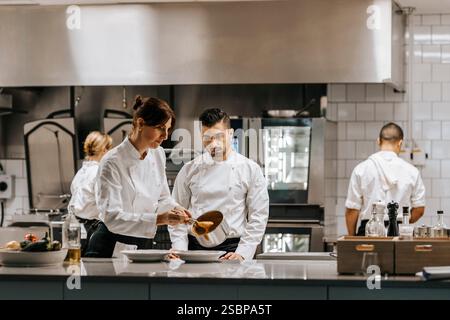 Lo chef maschile guarda la collega che versa la salsa nel piatto mentre si trova vicino al bancone della cucina del ristorante Foto Stock