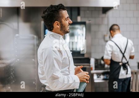 Vista laterale del giovane chef maschile felice in uniforme bianca in piedi presso la cucina del ristorante Foto Stock