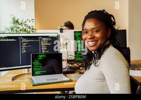 Ritratto di una giovane donna felice professionista IT seduta vicino a un PC desktop e a un notebook nell'ufficio tecnico Foto Stock