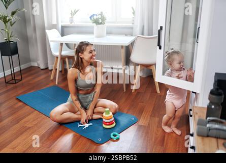 Una madre felice e gioiosa sta praticando la sua routine yoga a casa, mentre il suo adorabile bambino gioca nelle vicinanze, mostrando una splendida combinazione di fit Foto Stock