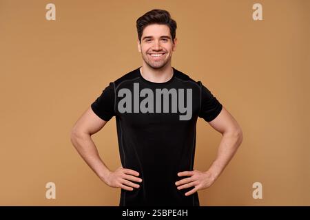 Un giovane allegro, vestito da elegante abbigliamento sportivo nero, si erge con sicurezza con le mani sui fianchi, sorridendo ampiamente contro un dorso caldo e invitante Foto Stock