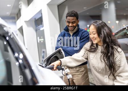 Una coppia multietnica felice che carica la sua nuova auto elettrica presso uno showroom della concessionaria, adottando uno stile di vita sostenibile e soluzioni energetiche ecologiche Foto Stock