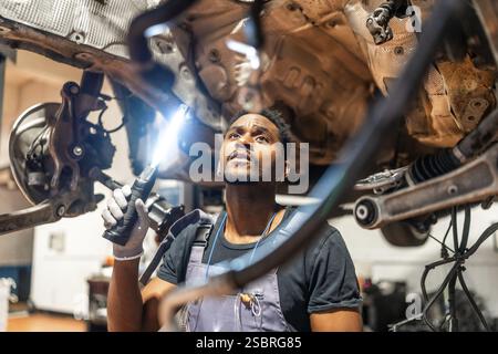 Giovane meccanico che utilizza una torcia elettrica, ispeziona la parte inferiore della vettura in un'officina professionale, concentrandosi sulla manutenzione e la riparazione Foto Stock