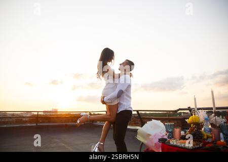 Una coppia gode di un momento romantico sul tetto mentre il sole tramonta. La donna, che indossa un vestito bianco, è sollevata di gioia dal suo partner. Una splendida arran Foto Stock