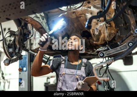 Giovane meccanico che utilizza la torcia e tiene in mano gli appunti, ispeziona il sottocarro di un'officina professionale Foto Stock