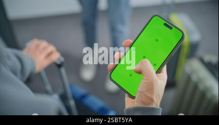 Terminal dell'aeroporto: Primo piano dell'uomo in piedi con valigetta, smartphone con mascherina verde Chroma Key display Mock Up, viaggio in vacanza. Modello video con segnaposto per i contenuti dei social media. Foto Stock