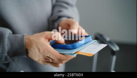Terminal dell'aeroporto: Primo piano di un uomo irriconoscibile che tiene in mano documenti con il biglietto, controlla le informazioni sul volo tramite smartphone, naviga su Internet o social network. Viaggiatore in viaggio. Foto Stock