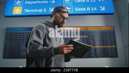 Terminal aeroportuale affollato: L'uomo con zaino utilizza un computer tablet e controlla la sua partenza sulla schermata Digital Flight Information. Un turista che va in vacanza. Visualizzazione degli orari aeroportuali in background. Foto Stock