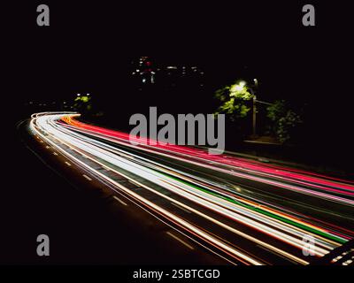 Un'autostrada curvilinea di notte, scattata con fotografie a lunga esposizione, che mostra strisce di luci colorate provenienti da veicoli di passaggio in un ambiente urbano. Foto Stock