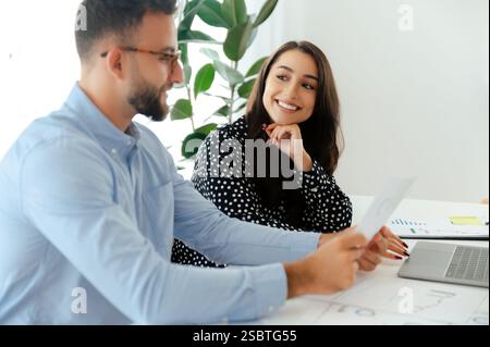 Uomo e donna professionisti motivati che discutono di lavoro presso una moderna scrivania, esaminano grafici e documenti finanziari con attenzione e interessi, sviluppano nuove idee e strategie, sorridono Foto Stock