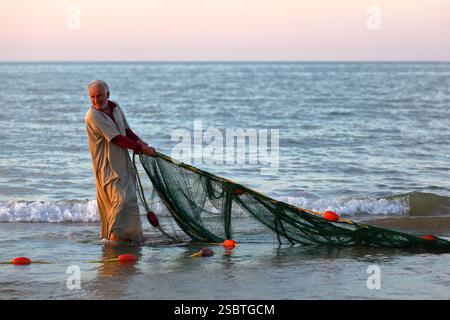 QURUM BEACH, MASCATE, OMAN - 28 DICEMBRE 2024: Pescatore omanita di ritorno dalla pesca e lavoro sulla sua rete da pesca sulla spiaggia di Qurum Foto Stock