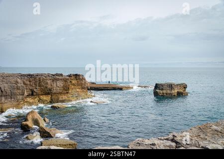 Una piccola insenatura costiera su Portland, Dorset, incorniciata da rocce e scogliere. In lontananza, un uomo solitario si erge su rocce sotto cieli luminosi e acque calme. Foto Stock