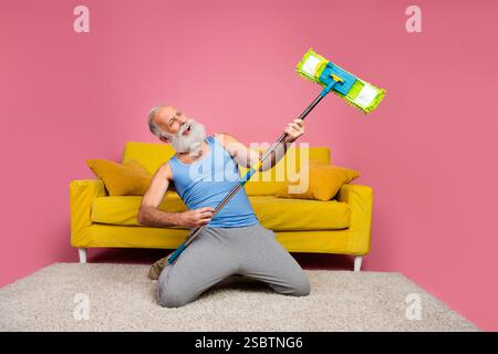 Uomo anziano in un abbigliamento casual che imita con passione la chitarra con un mop in un interno vivace Foto Stock