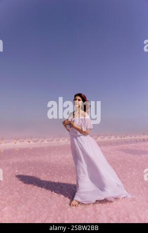 Woman White Dress Pink Lake - Una donna che indossa un abito bianco cammina sulla superficie di un lago rosa. Il lago è circondato da un cielo blu. Foto Stock