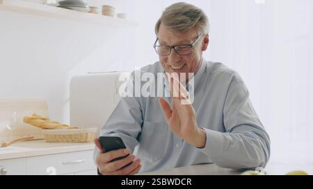 Pensionato sorridente mentre videochiama in una cucina luminosa Foto Stock