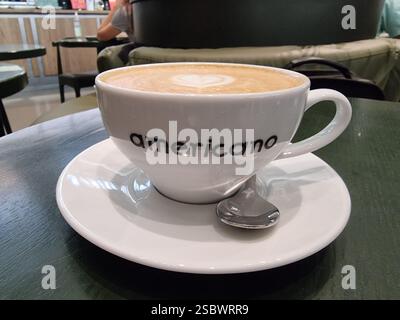 Tazza da caffè bianca luminosa con cappuccino, con testo americano (su un tavolo in un ristorante caffetteria) Foto Stock