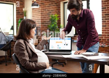 Uomo barbuto che sottolinea l'errore nell'analisi aziendale sul laptop, durante l'incontro con una donna bianca incinta. Colleghi di sesso maschile e femminile che utilizzano dispositivi digitali per discutere della strategia finanziaria dell'ufficio di startup. Foto Stock