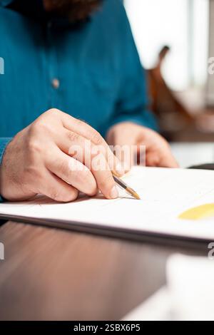Attenzione selettiva da parte del dipendente che tiene la penna sui documenti finanziari sul tavolo. Primo piano di una persona caucasica che scrive e rivede i dati delle ricerche aziendali negli appunti dell'ufficio startup. Foto Stock