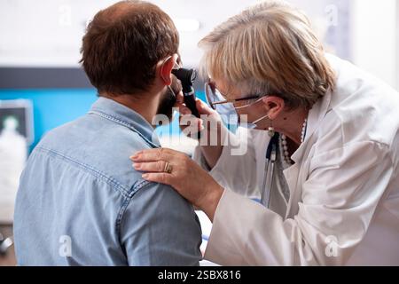 La donna in pensione esamina attentamente l'orecchio dell'uomo barbuto seduto in clinica, utilizzando strumenti professionali per controllare l'infezione. Vecchio medico con otoscopio che fa visita medica su paziente di sesso maschile. Foto Stock