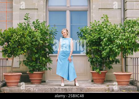 Ritratto a tutta lunghezza del corpo di una bella donna di mezza età di 50 -55 anni, con indosso un abito blu, che posa all'esterno accanto a alberi di limone piantati Foto Stock