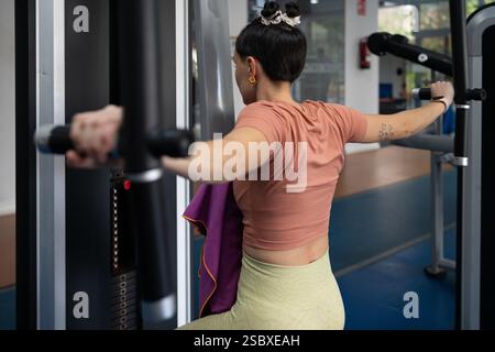 Giovane donna sportiva che allena i muscoli della schiena usando una macchina per pesi in palestra, mostrando impegno e dedizione agli obiettivi di fitness, con un asciugamano viola sulla schiena Foto Stock